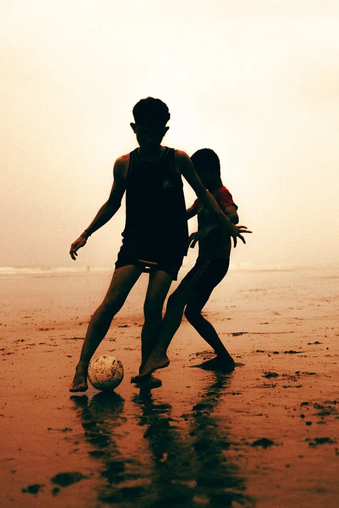 Two children playing soccer on the beach at sunset, one with a ball at their feet and the other trying to kick or reach the ball.