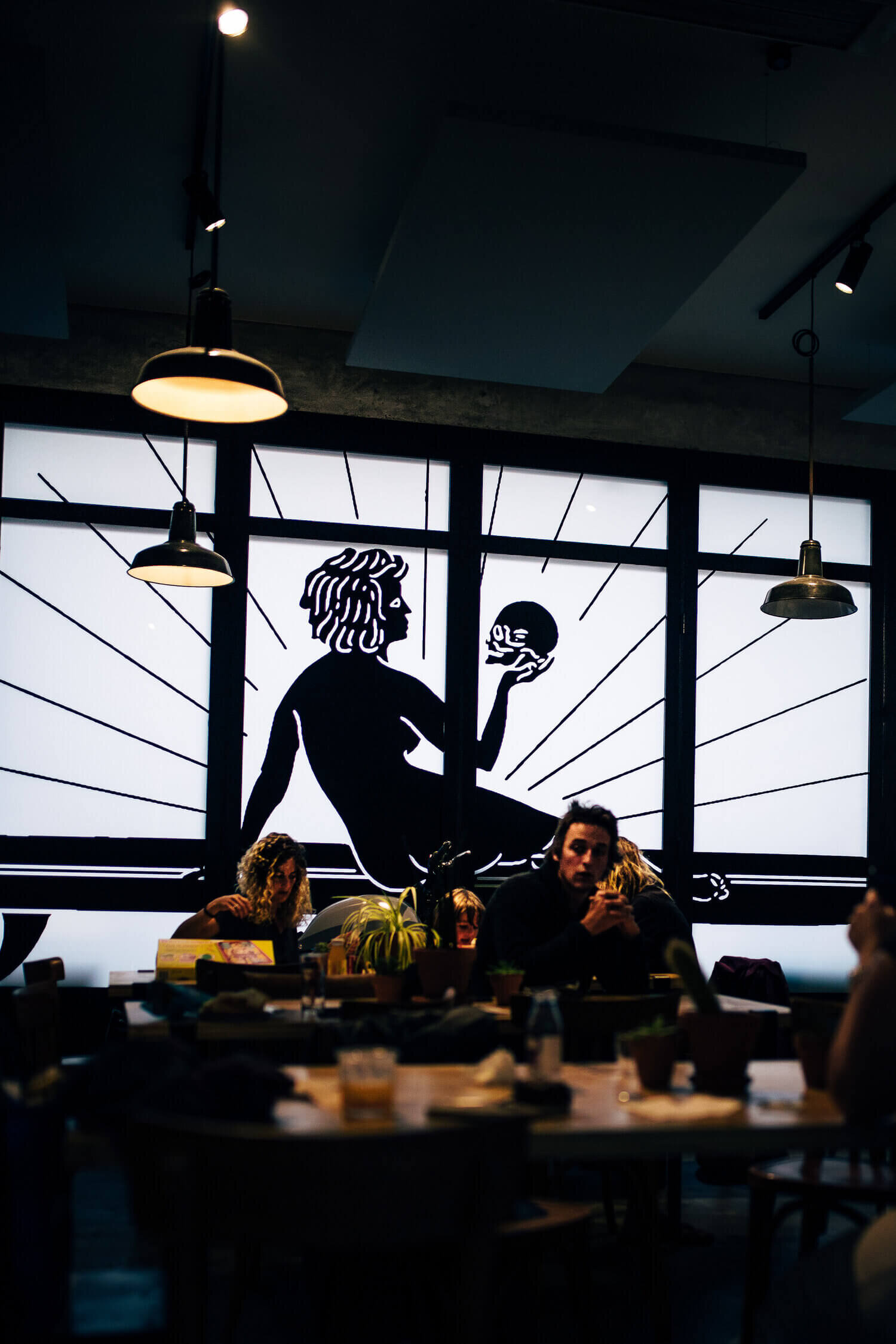 Intérieur d'un café avec des clients assis, une grande illustration d'une silhouette tenant un crâne est visible sur la fenêtre en arrière-plan, et des lampes suspendues éclairent la pièce.