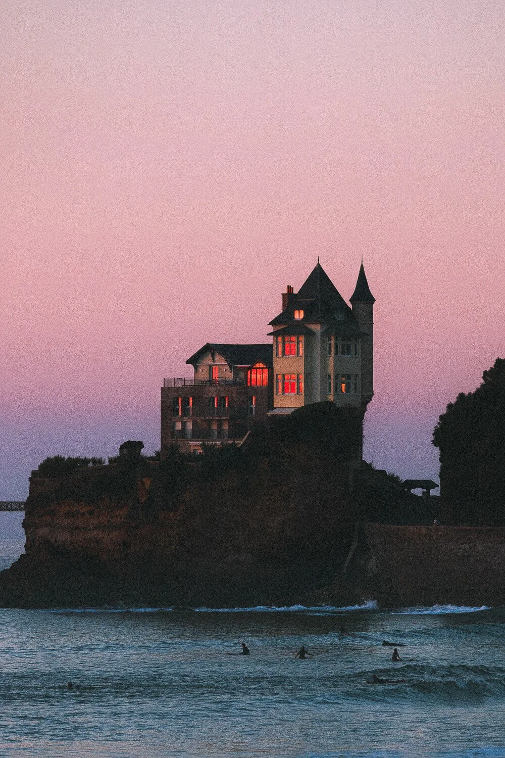 A historic mansion on a cliff overlooking the ocean at sunset, with surfers in the water.