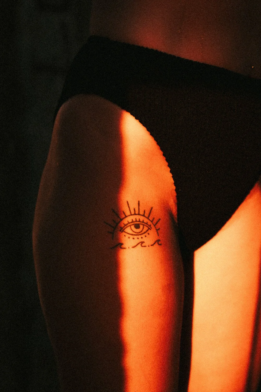 Tattoo of an eye with extended eyelashes above a wave on a person's upper thigh, illuminated by warm orange light.