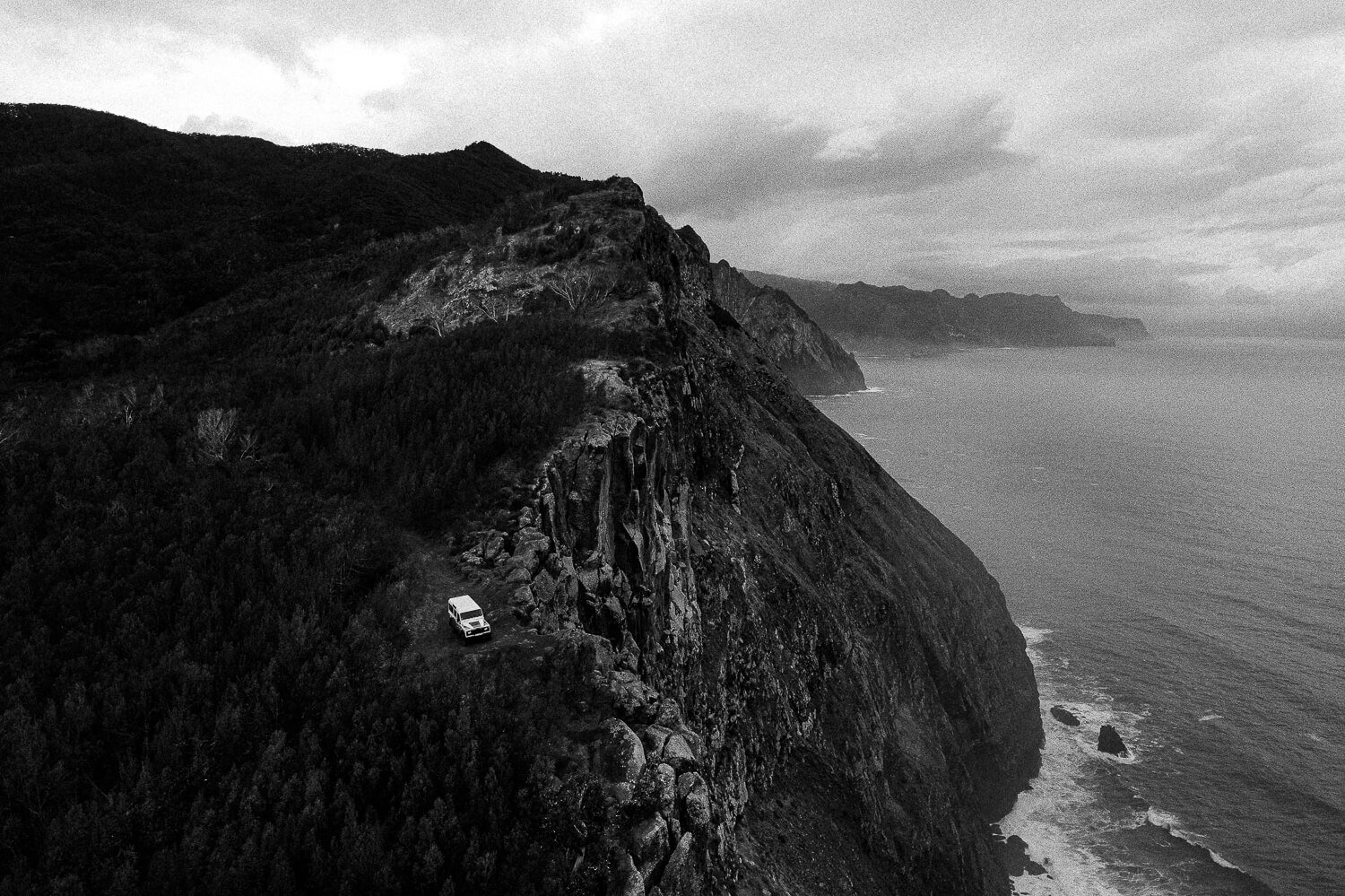 A black and white aerial photograph of a steep coastal cliff with dense vegetation, overlooking the ocean with visible waves, and a small vehicle parked on the rocky terrain near the cliff's edge.