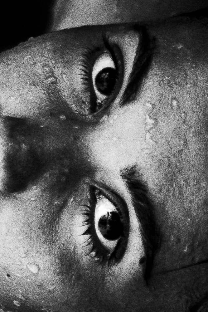 Close-up of a person's face with water droplets, emphasizing the eyes, in black and white.