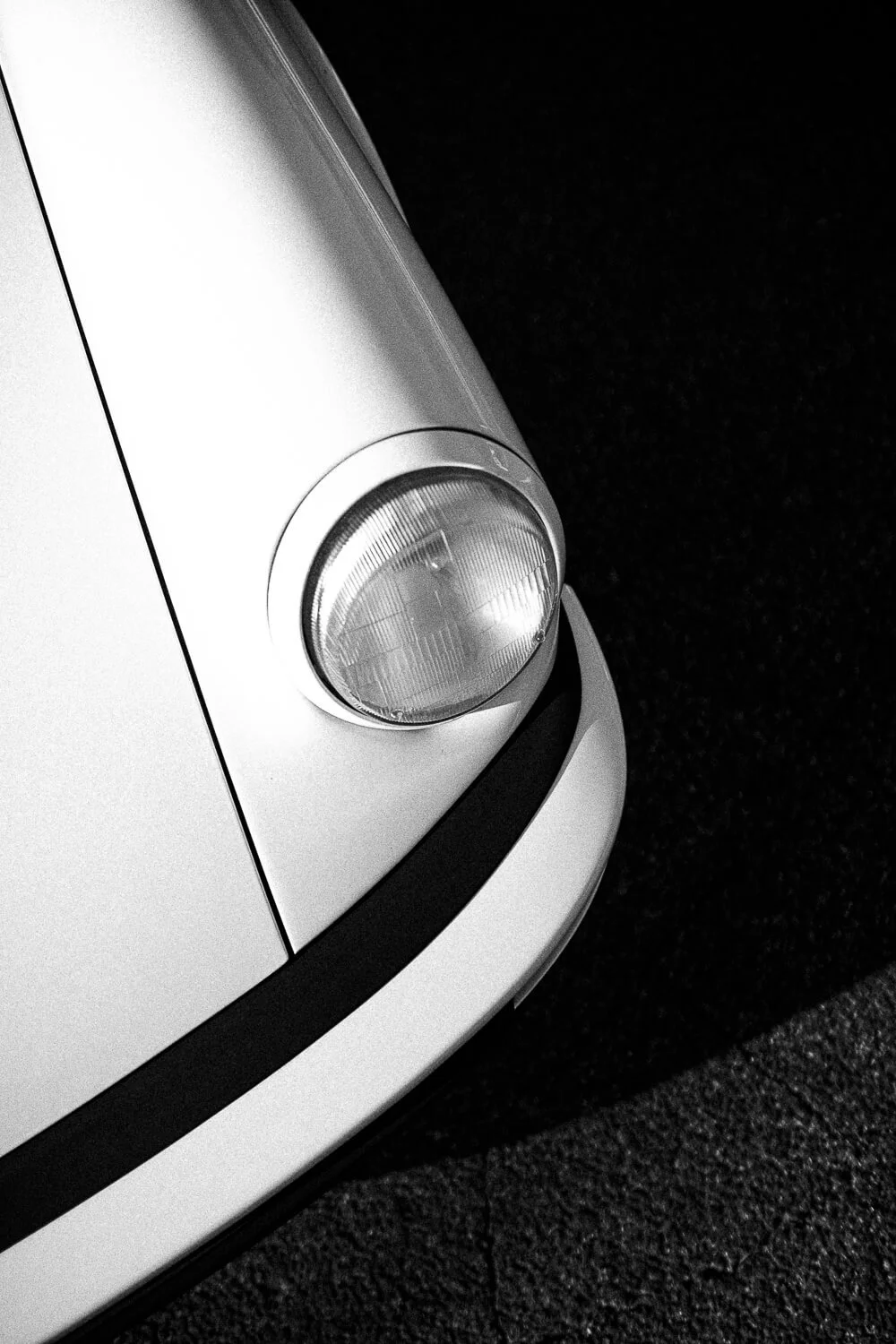Close-up of the front left corner of a classic white Volkswagen Beetle car, focusing on the round headlight and black bumper detail, on a dark textured surface.