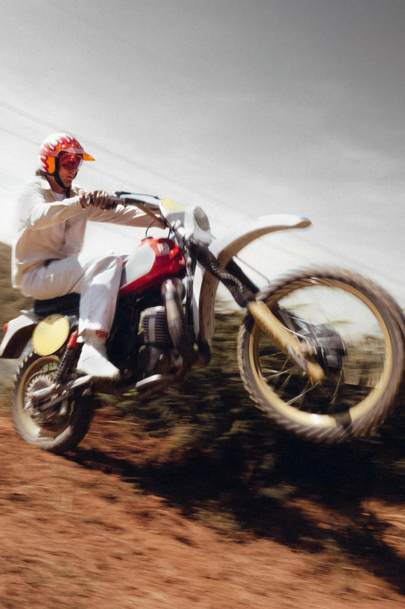 A person riding a dirt bike on a dirt track during daytime, wearing a red helmet with flames design and glasses, with a blurred background.