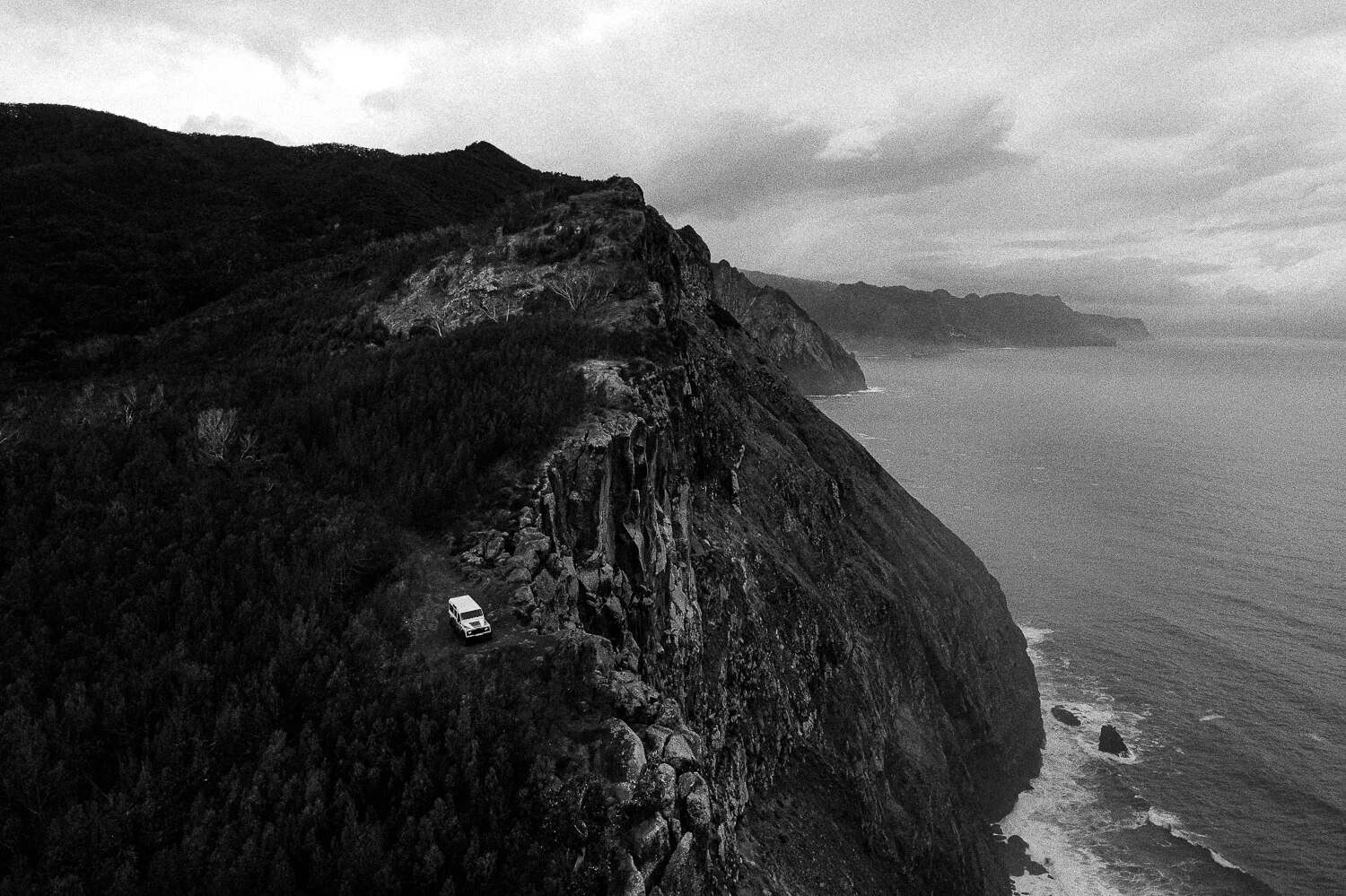 A black and white photo of a rugged coastline with steep cliffs overlooking the ocean, and a white vehicle parked on a narrow dirt path on the cliffside.