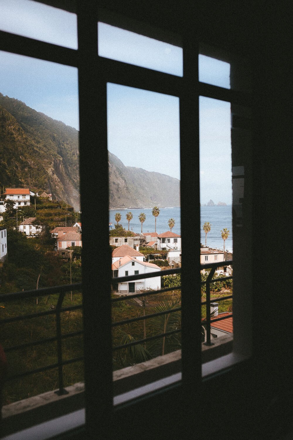 View through a window with black bars showing a coastal town with white houses, palm trees, green hills, and the ocean under a clear sky.