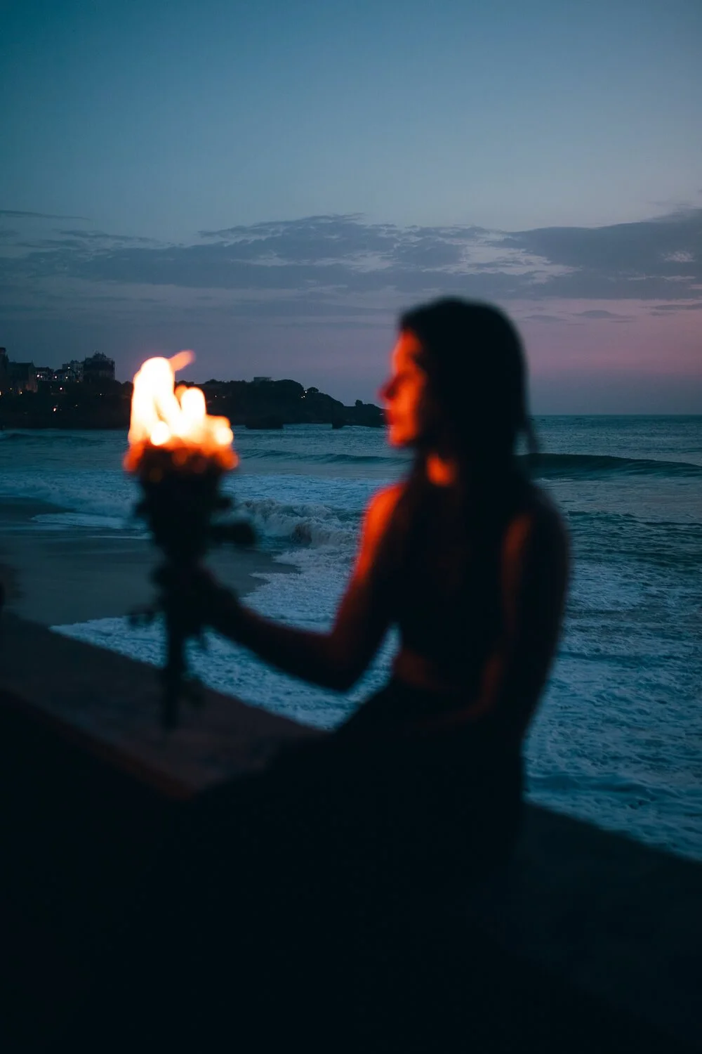 A woman on the beach at sunset holding a torch with a small flame, with the ocean and a distant shoreline in the background.