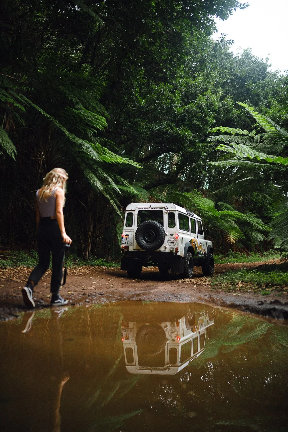 A woman walking with a camera near a white off-road vehicle parked on a dirt trail surrounded by lush green foliage in a jungle or forest.