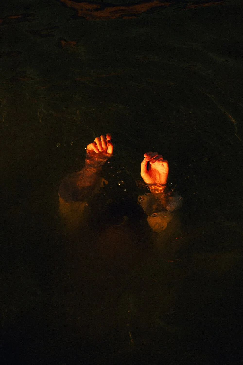 Hands and head of a person submerged in dark water, with fists raised above the surface in a night scene.