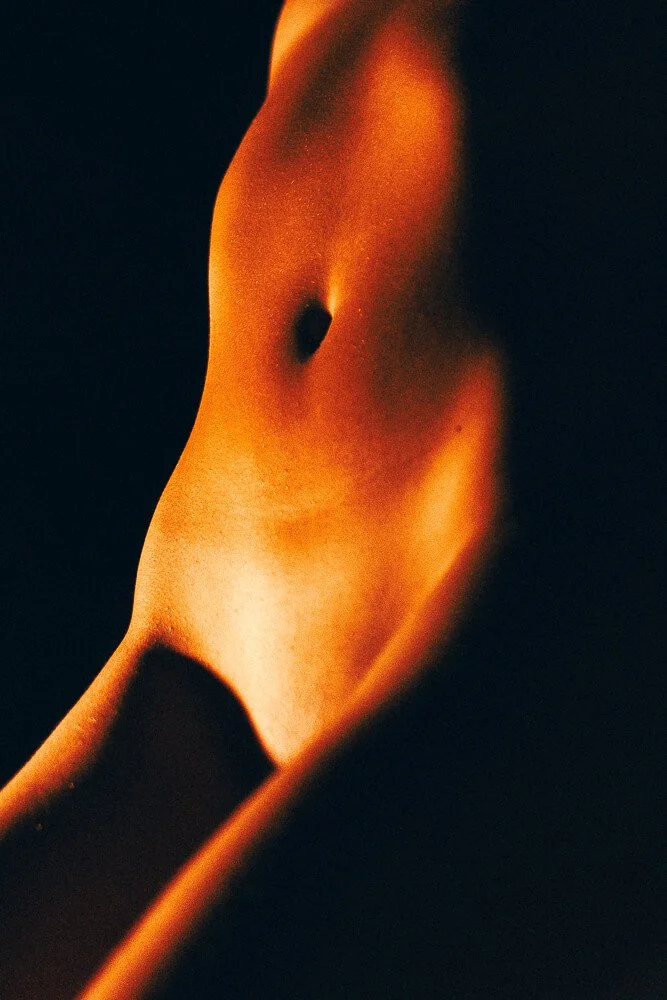 Close-up of a person's bare midriff with a focus on the navel, illuminated with warm, soft lighting against a dark background.