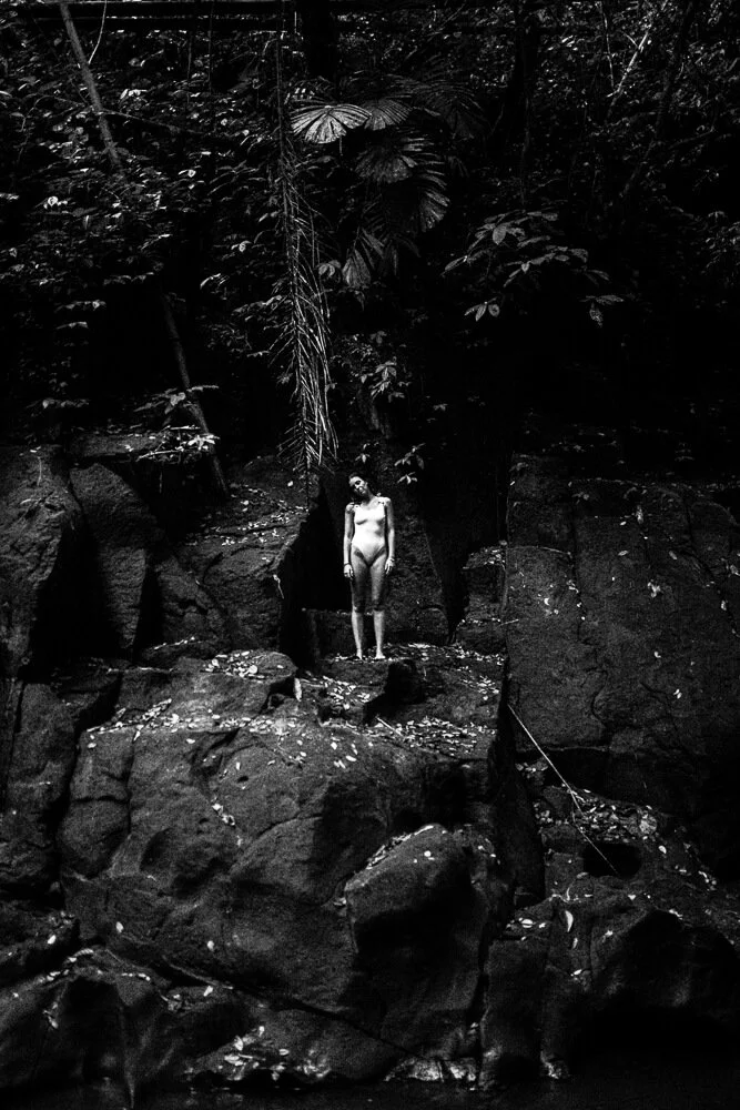 A black and white photo of a woman standing nude on rocks in a dense forest, surrounded by large trees and foliage.