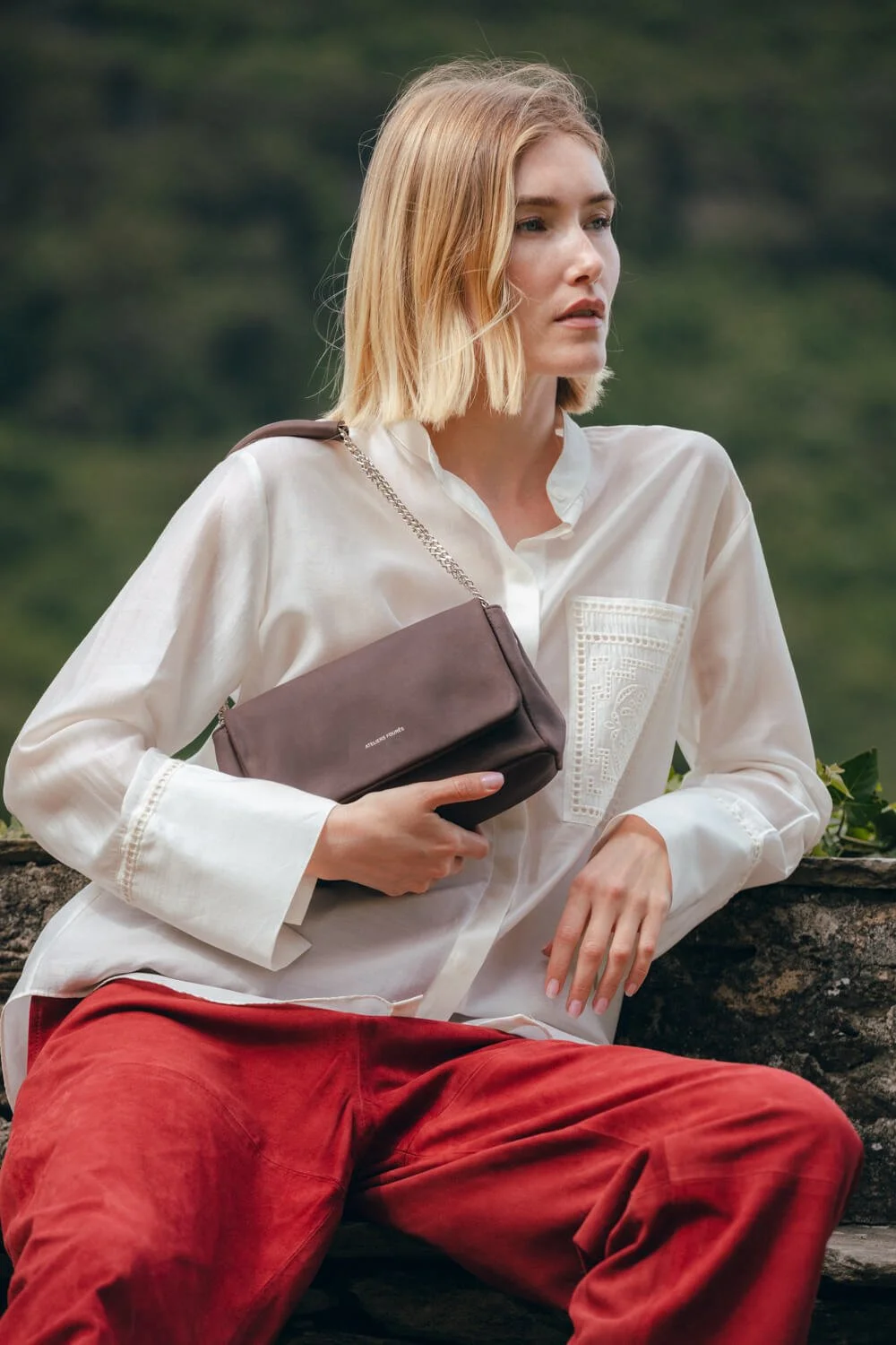 A young woman with blonde hair in a white blouse and red pants sitting outdoors on a log, holding a brown shoulder bag.