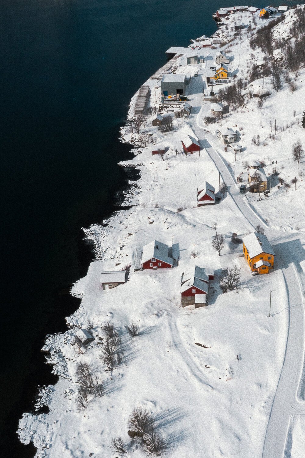 Paysage hivernal montrant des maisons colorées le long d'une côte enneigée, avec une mer sombre à gauche et une route enneigée à droite.