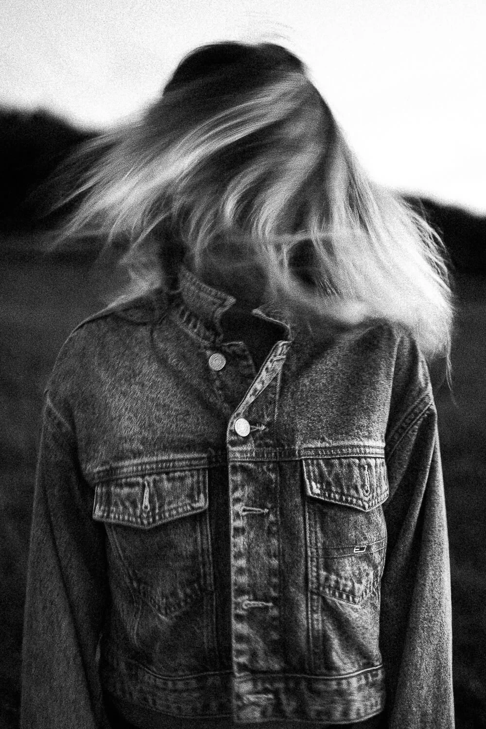 Black and white photo of a person wearing a denim jacket with their head down and hair covering their face, outdoors at dusk or night.