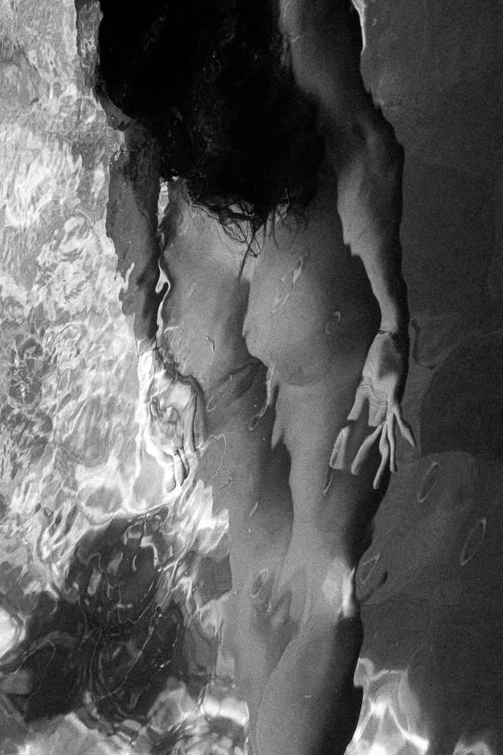 A woman with curly hair lies in shallow water at the beach, with her arm resting on the wet sand and her head turned away from the camera.
