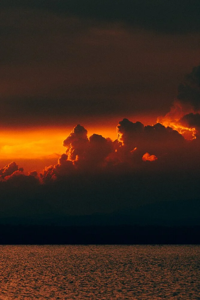 Sunset over a body of water with dark clouds and a fiery orange sky.