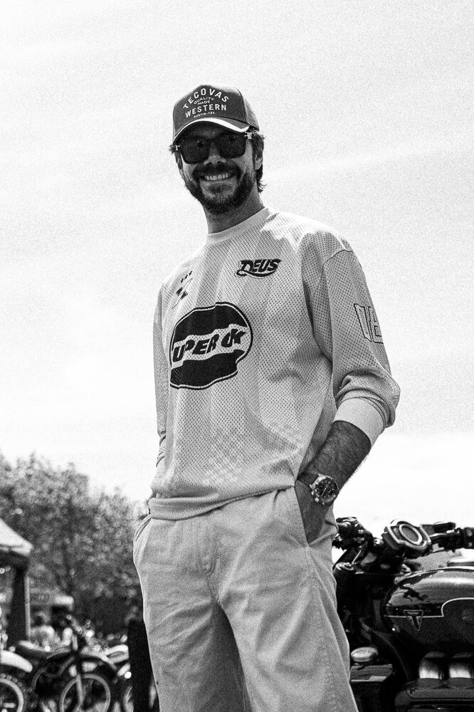 A man standing outdoors wearing sunglasses, a baseball cap, a long-sleeve sports shirt with logos, and light-colored pants. He is smiling and has one hand in his pocket, with motorcycles or bicycles in the background.