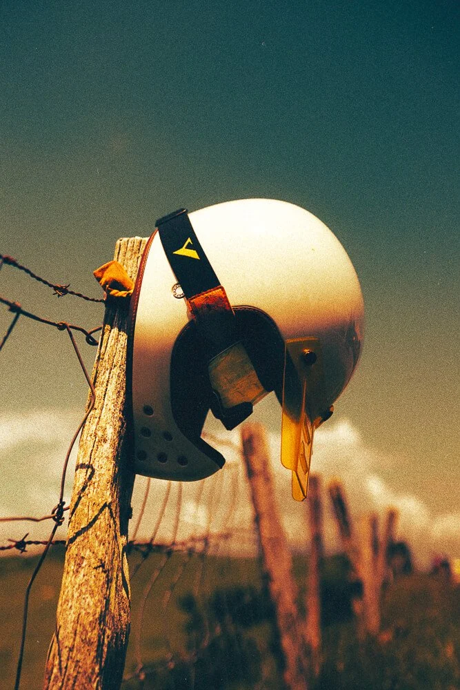 A white motorcycle helmet hanging on a wooden fence post with barbed wire, set outdoors during sunset or sunrise.