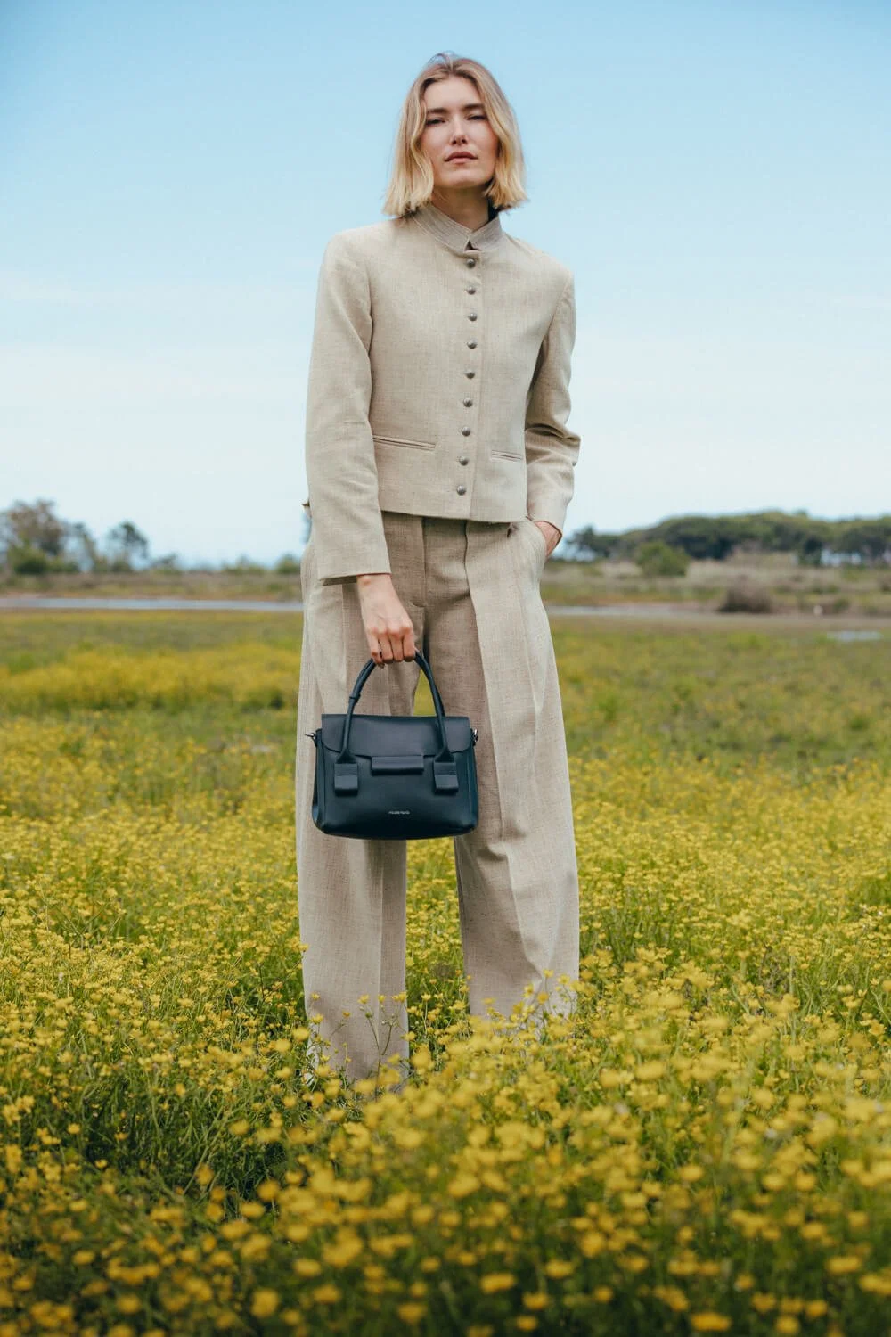 A woman in a beige suit holding a black handbag stands in a field of yellow flowers with a clear sky in the background.