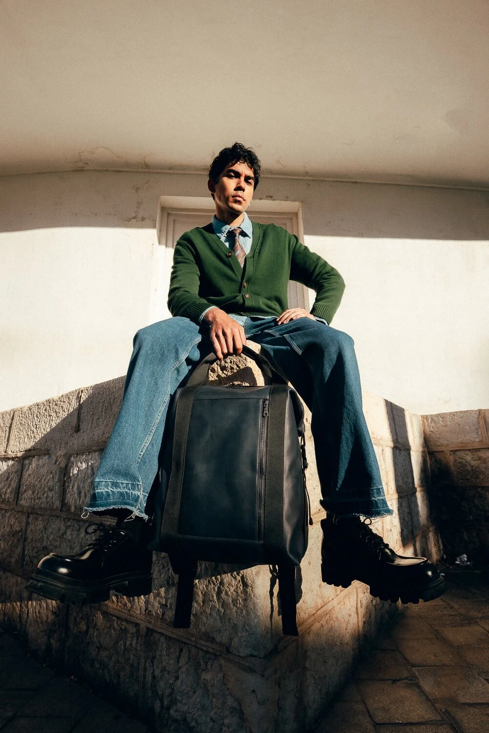 A man sitting on a stone ledge with a black backpack in front of a white wall, wearing a green cardigan, blue jeans, black boots, and a tie, with his right hand resting on the backpack.
