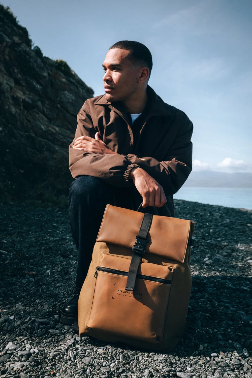 A man in a brown jacket kneels on a rocky beach with a large tan backpack in front of him. He is looking to the side with folded arms, near a cliff with a cloudy sky in the background.