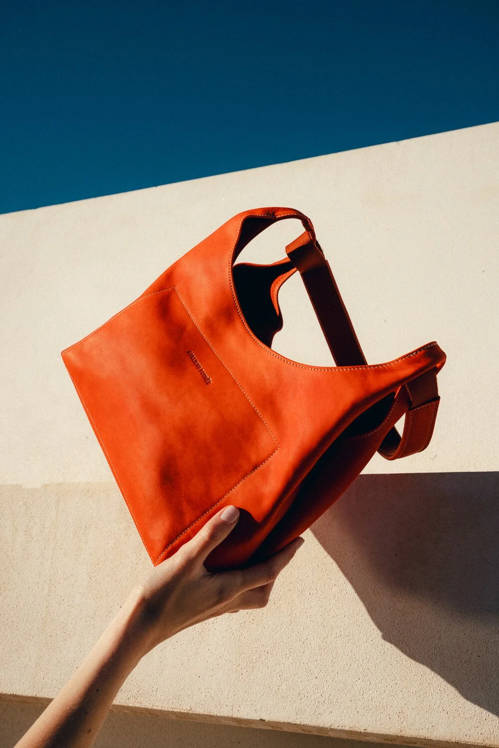 A person holding an orange leather handbag with a minimalist design, against a background of cream-colored surface and a blue sky.