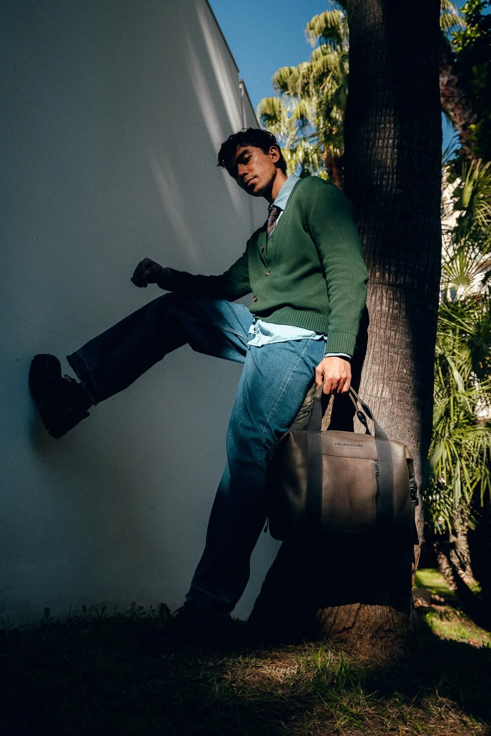 A man leaning against a tree with one foot on a white wall, holding a black duffel bag, wearing a green cardigan and jeans.