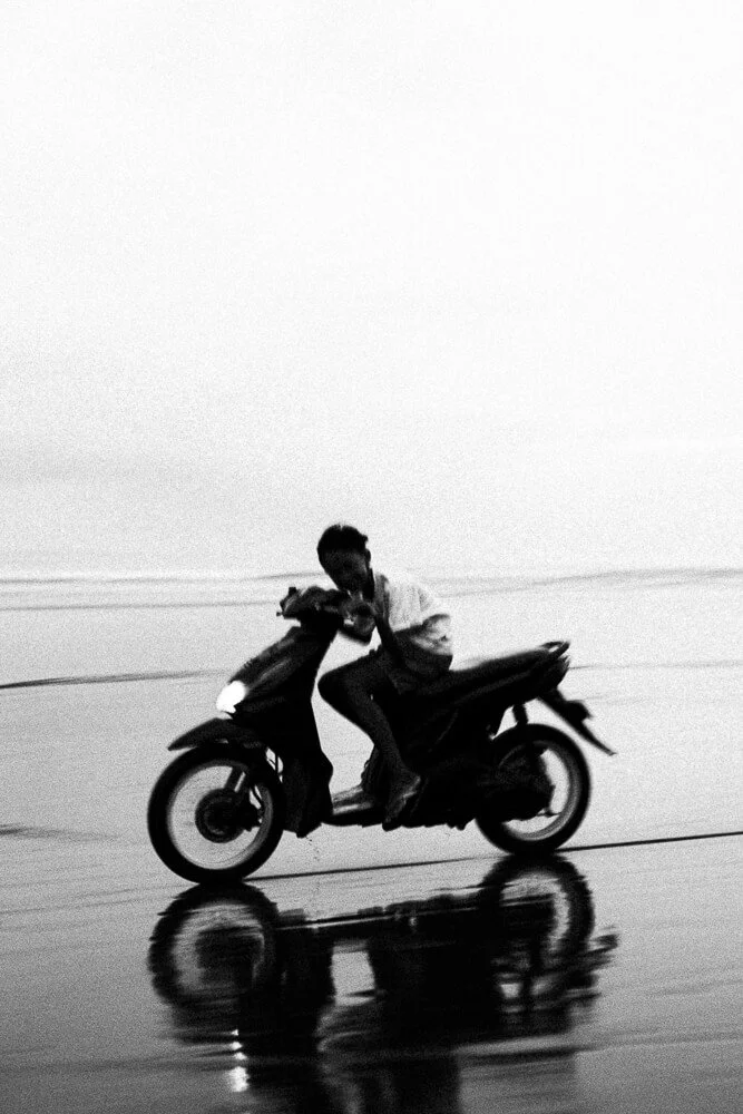 A person riding a scooter on a wet surface, creating a reflection of the scooter and rider. The scene appears to be outdoors, possibly near a beach or open area.