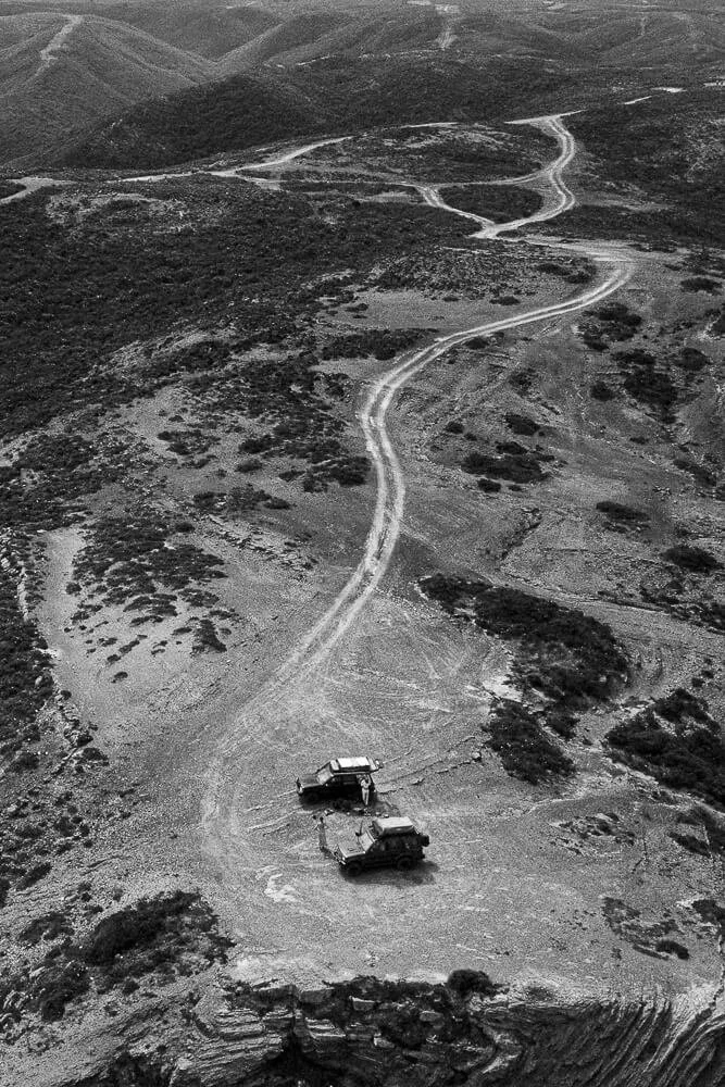 Two off-road vehicles parked on a winding dirt trail in a mountainous, arid landscape with sparse vegetation.