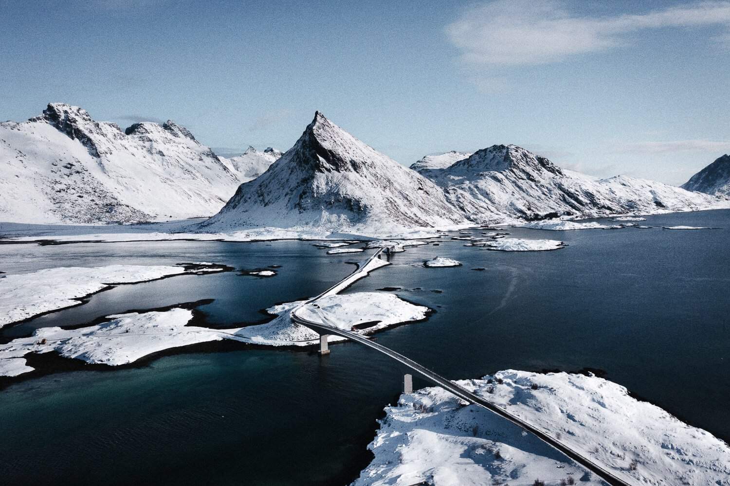 Paysage glaciaire avec montagnes enneigées, un pont suspendu au-dessus de l'eau, et des îles recouvertes de neige.
