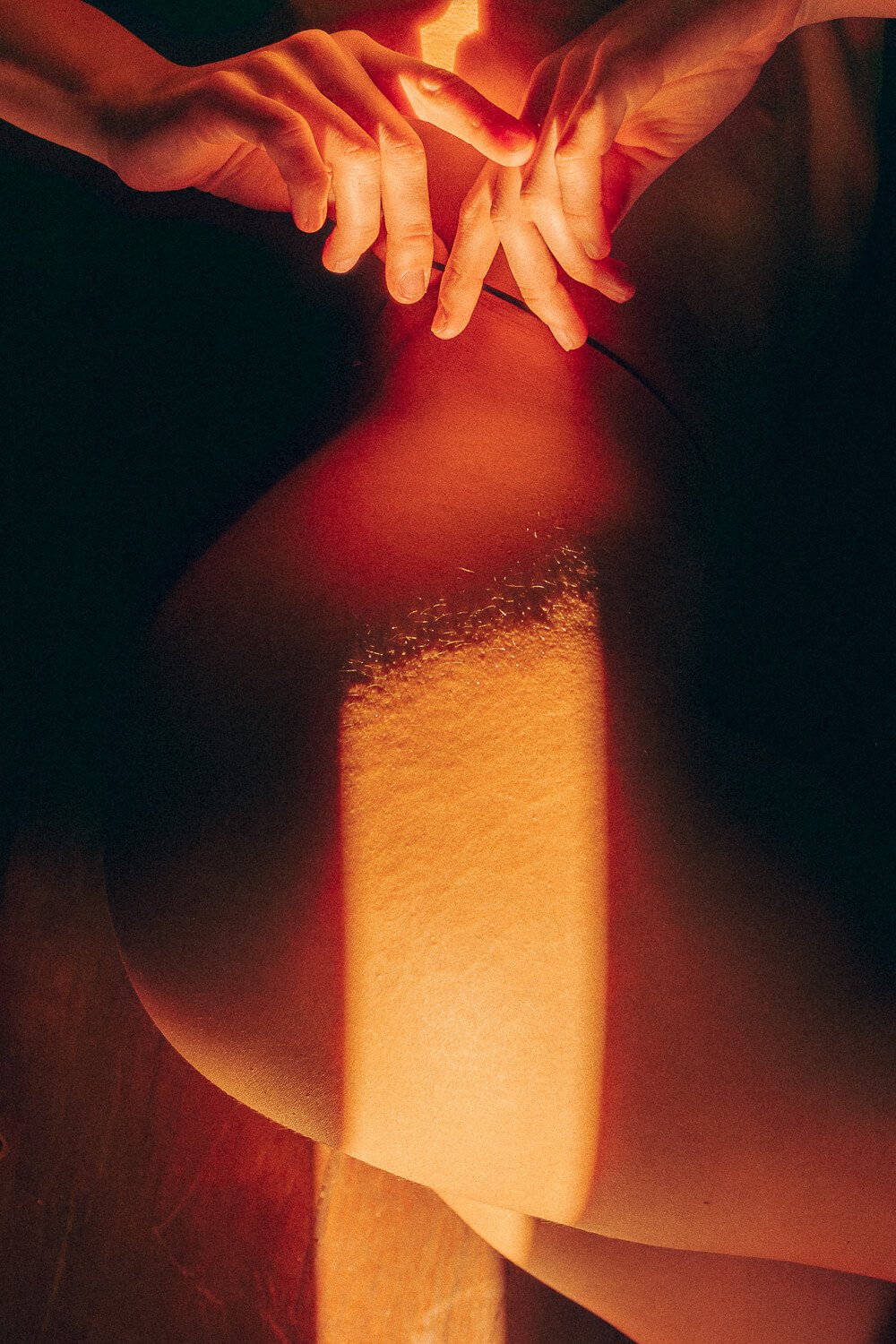 Close-up of two hands clasped together resting on a thigh, illuminated by soft warm lighting highlighting skin texture and muscle contours.