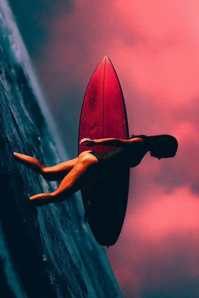 A person standing on a surfboard on the ocean, with a colorful pink and purple sky in the background.