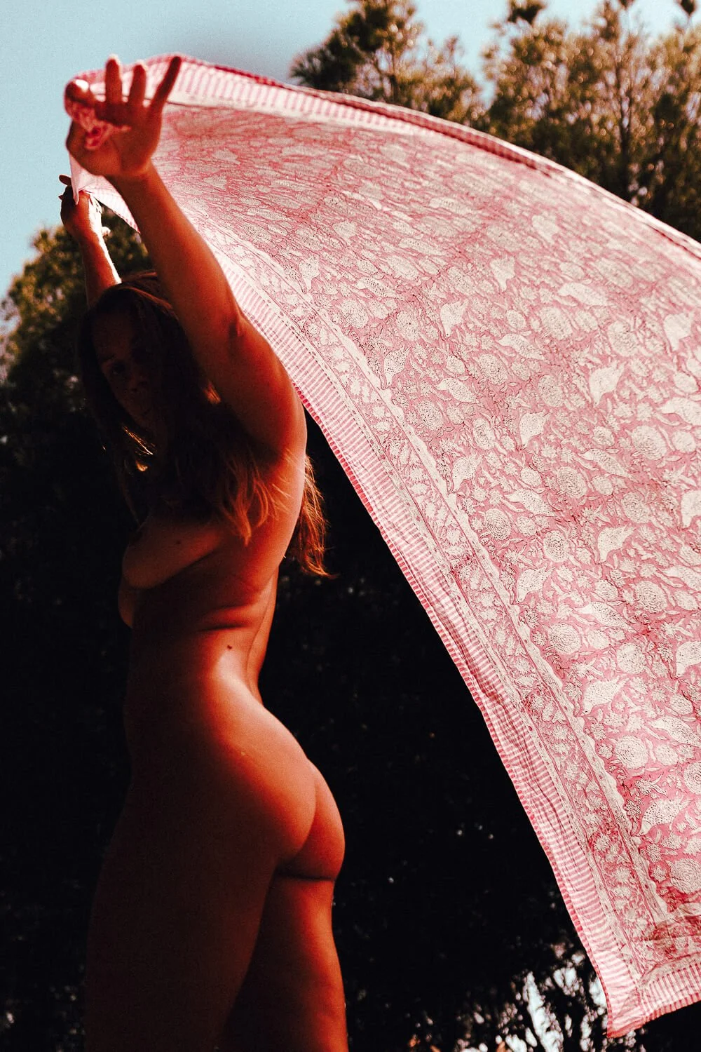A nude woman with long hair holding a pink lace umbrella outdoors during sunset.