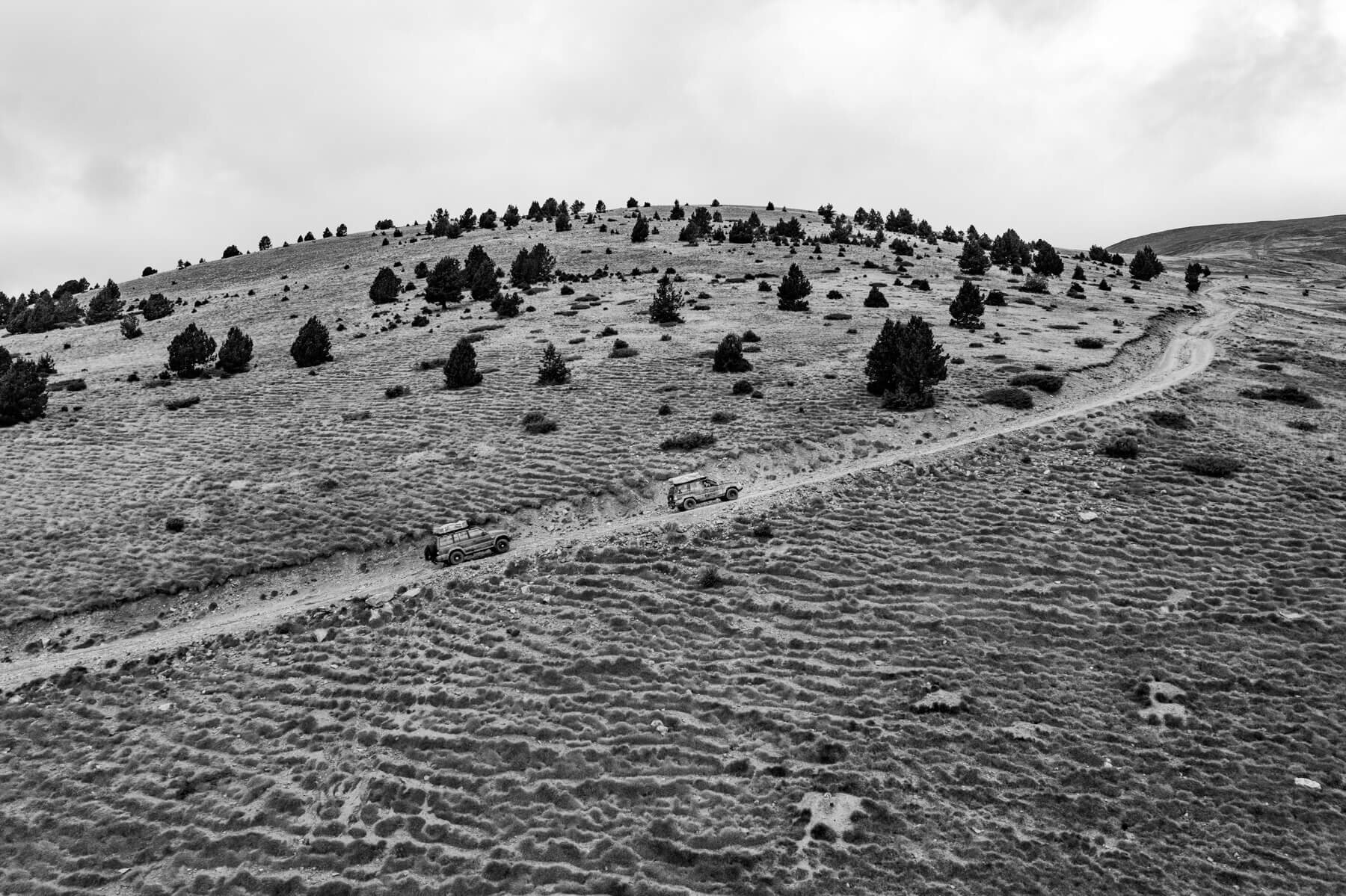 Deux véhicules tout-terrain sur un chemin de montagne avec des buissons dispersés et un ciel nuageux.