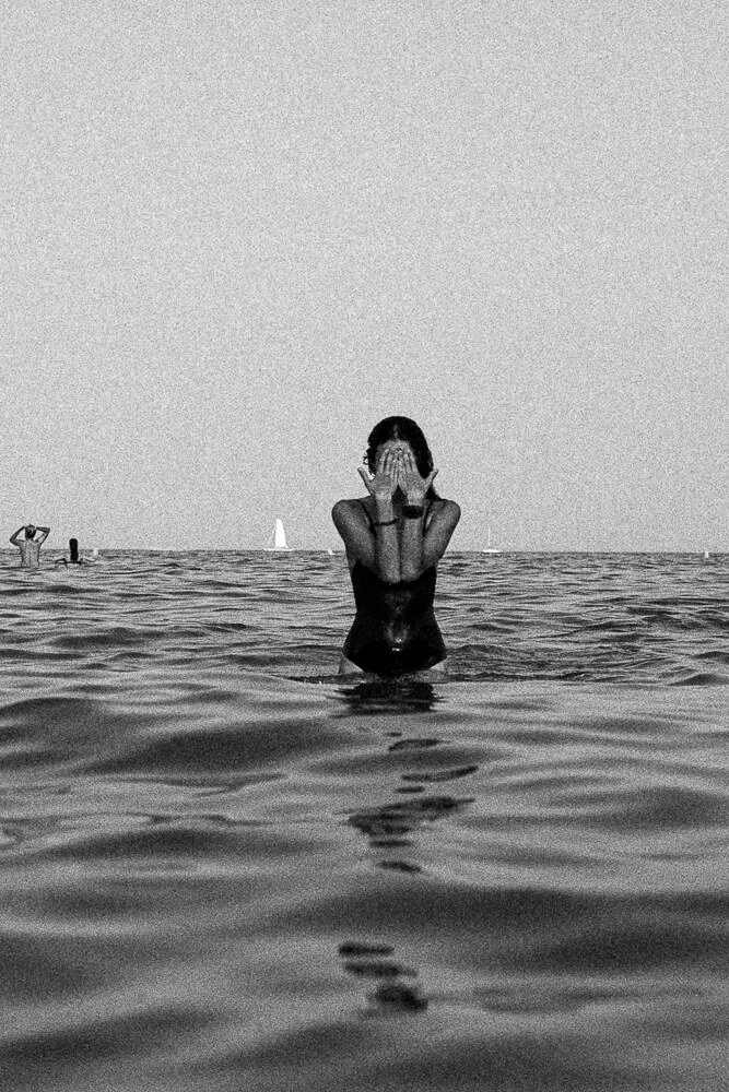 A woman standing in the ocean with her hands covering her face, surrounded by water, with sailboats in the background, in black and white.