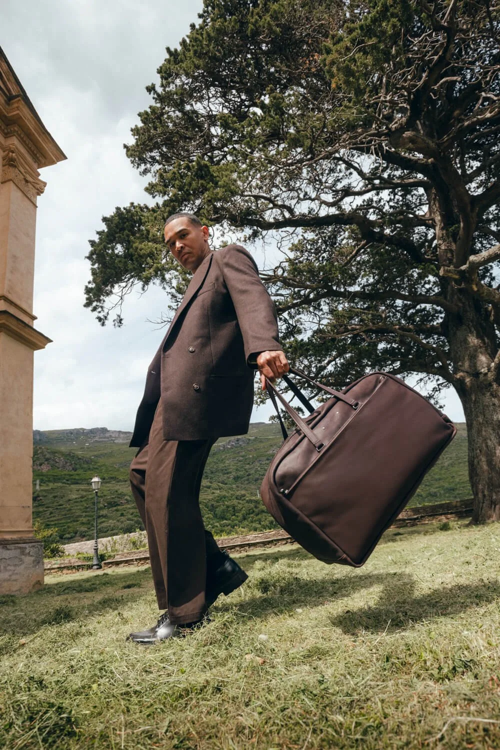 A man in a brown suit carrying a large brown suitcase outdoors, near a large tree with a scenic background of green hills and cloudy sky.