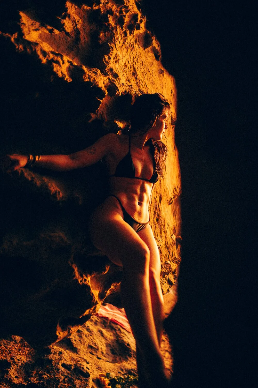 A woman in a black bikini lying on a sandy beach at sunset, with the sun casting an orange glow on her body and the sand behind her.