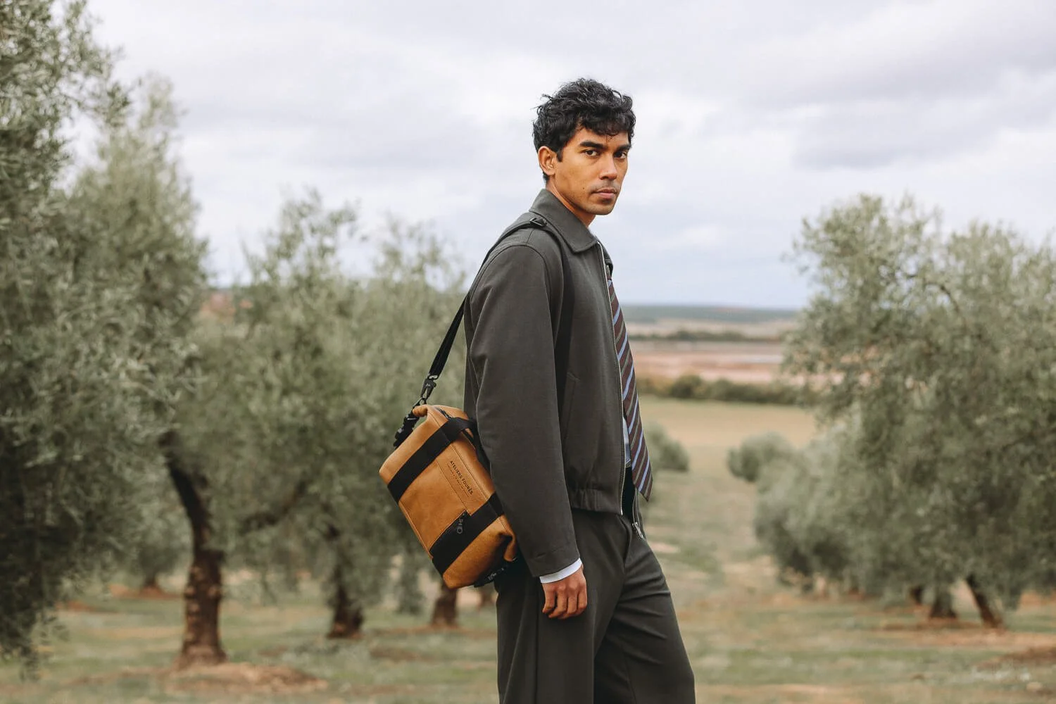 A man in a dark jacket and tie walking through an olive grove with a tan shoulder bag.