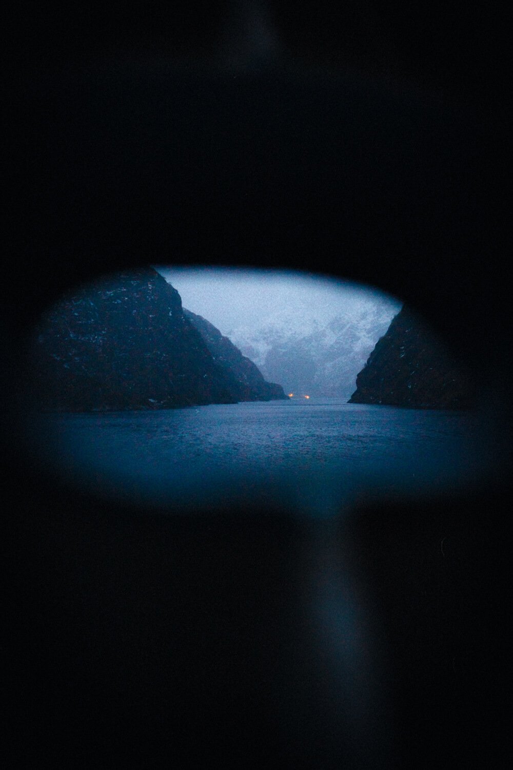 Vue d'un fjord à travers un trou dans une roche ou un tunnel, avec des montagnes enneigées en arrière-plan et un peu de lumière