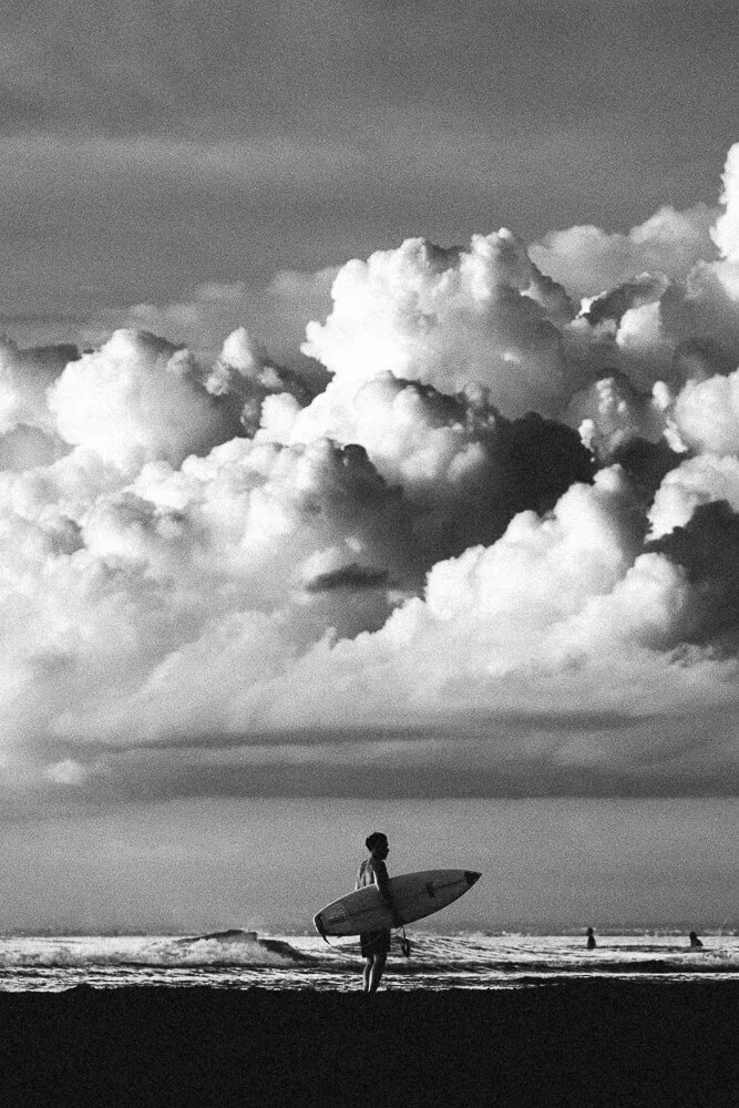 A person standing on the beach holding a surfboard, with a cloudy sky and others surfing in the background.