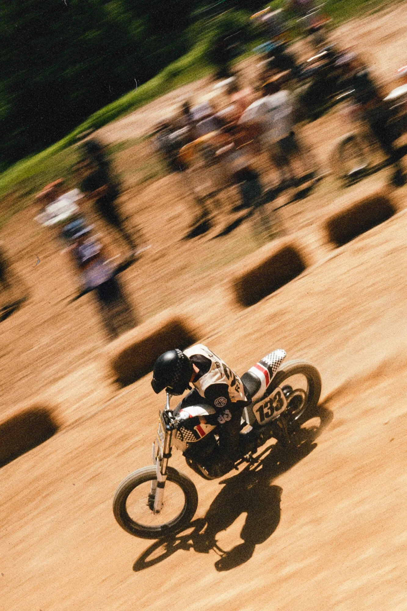 A dirt bike rider wearing a black helmet and racing gear riding on a dirt track with a dirt bike numbered 133, with blurred spectators and trees in the background.