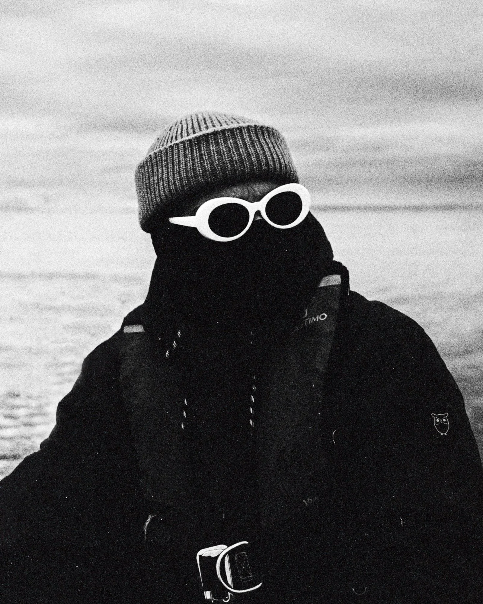 Personne en vêtements d'hiver portant un bonnet, des lunettes de soleil rondes, et un cache-cou, dans un paysage enneigé.