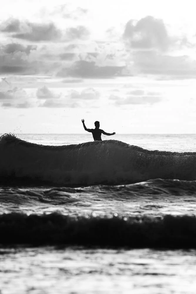 A person stands on a surfboard in the ocean, raising one hand while waves crash around them at sunset or sunrise.