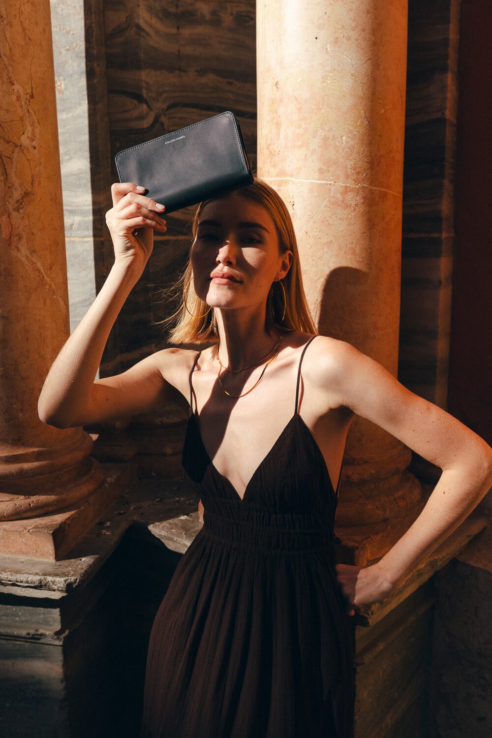 A woman in a black spaghetti strap dress is standing in front of large stone columns, holding a black wallet above her forehead, with sunlight casting shadows on her face and body.