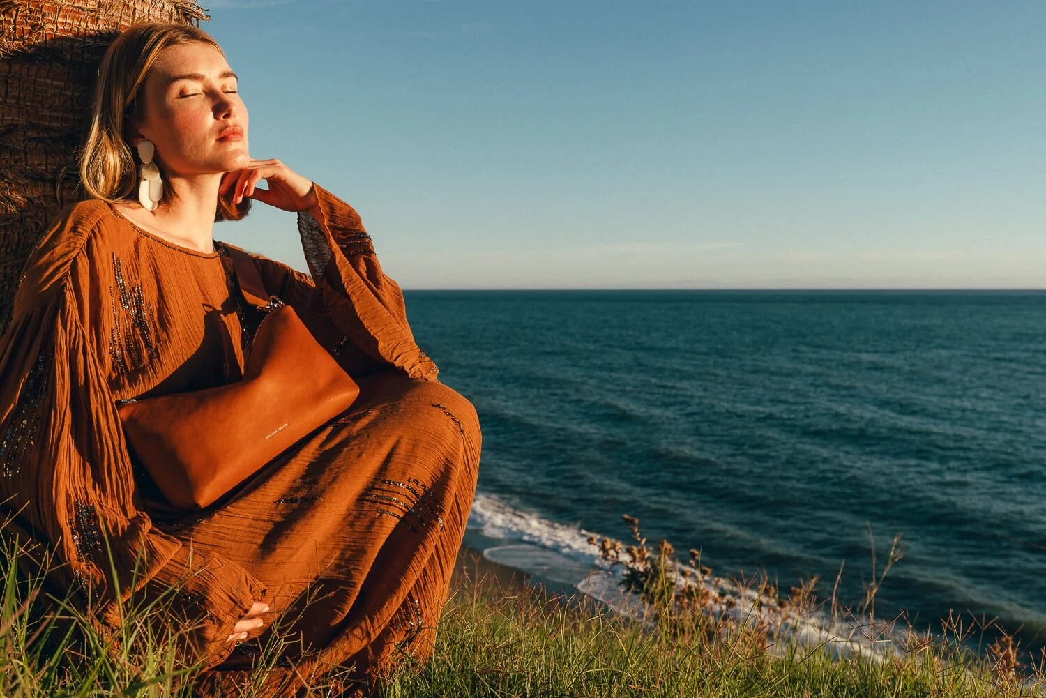Woman in an orange dress sitting on grass by the ocean, eyes closed, facing the sunset with a tan handbag on her lap.