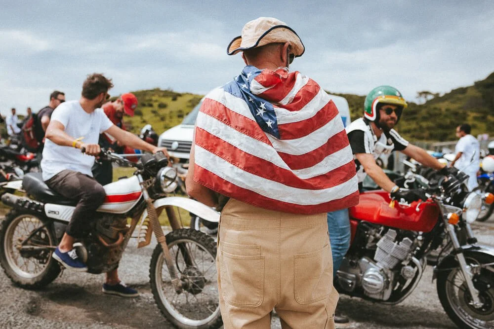 Person wrapped in an American flag standing among motorcyclists at an outdoor gathering.