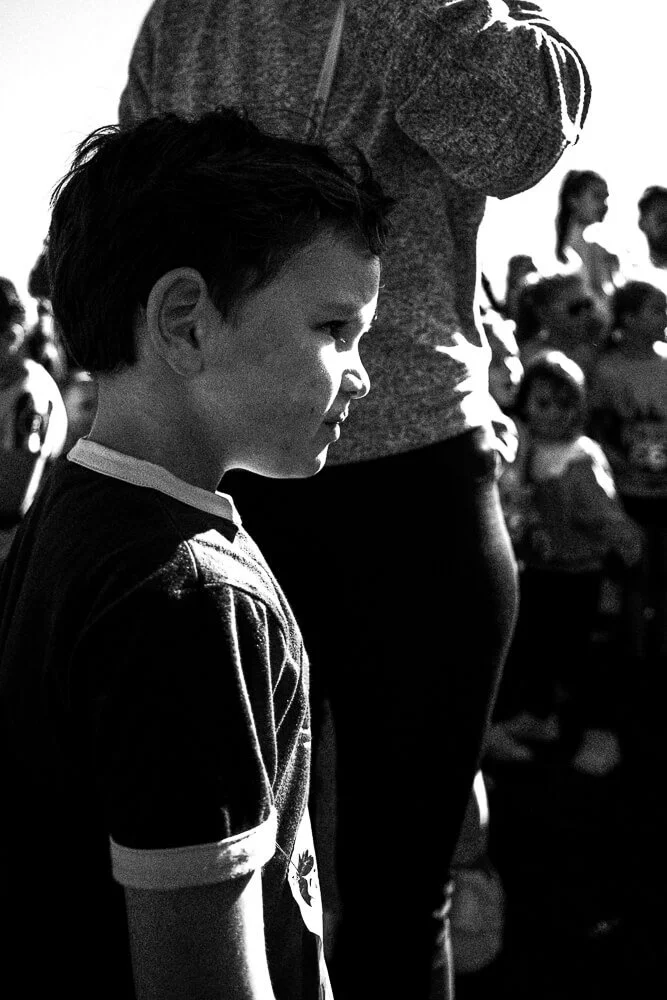 A young boy standing in profile, surrounded by a crowd of people, some children, in an outdoor setting during daytime.