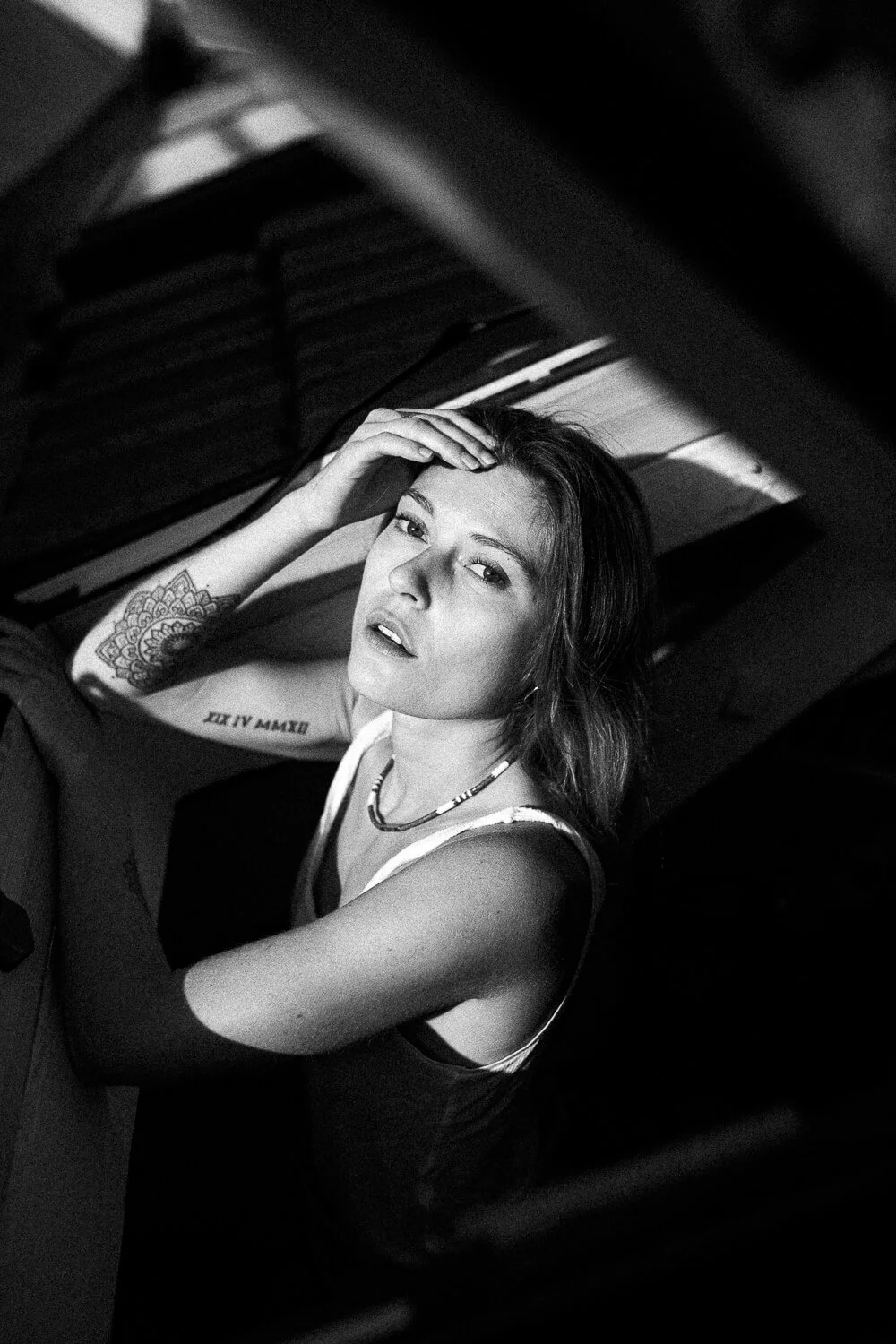 A young woman with tattoos and a necklace, sitting on the floor next to a piano, looking at the camera with her hand touching her head, black and white photo.
