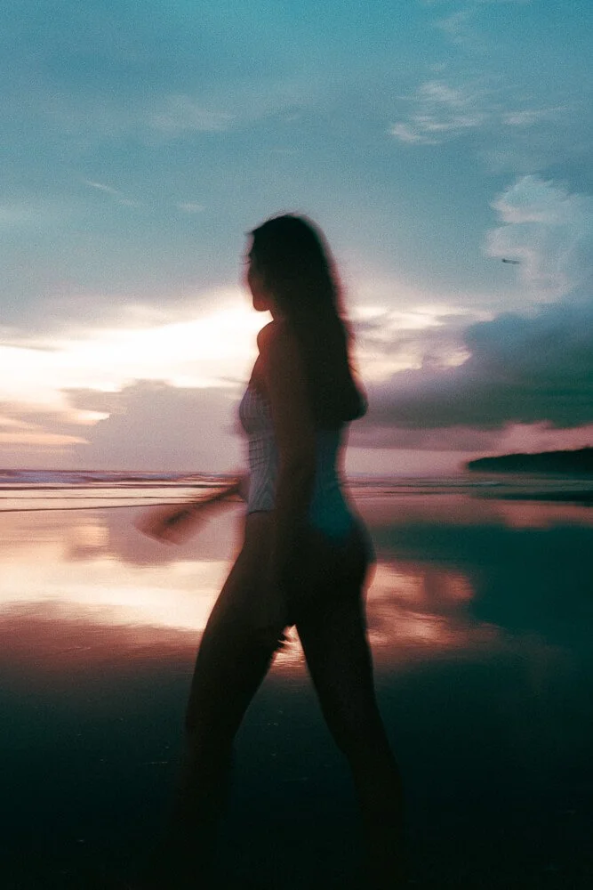 Silhouette of a woman walking along the beach at sunset or sunrise, with clouds and water reflecting the sky's colors.
