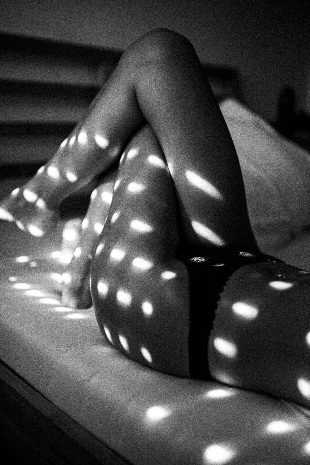 Black and white photo of a person's crossed legs on a bed, with light patterns of dots creating a polka-dot effect on the skin.